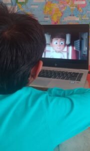 A child is sitting in front of a laptop, watching an animated movie. The screen displays a cartoon character with an expressive face. The child is wearing a bright blue shirt, and a world map is visible on the wall in the background. The setting suggests a fun and engaging learning or entertainment moment.