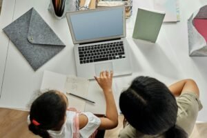A child and an adult learning together at a desk with a laptop, notebook, and stationery, fostering education and creativity.
