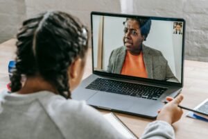 A young girl with braided hair attends an online class on her laptop, taking notes while listening to her teacher on a video call.